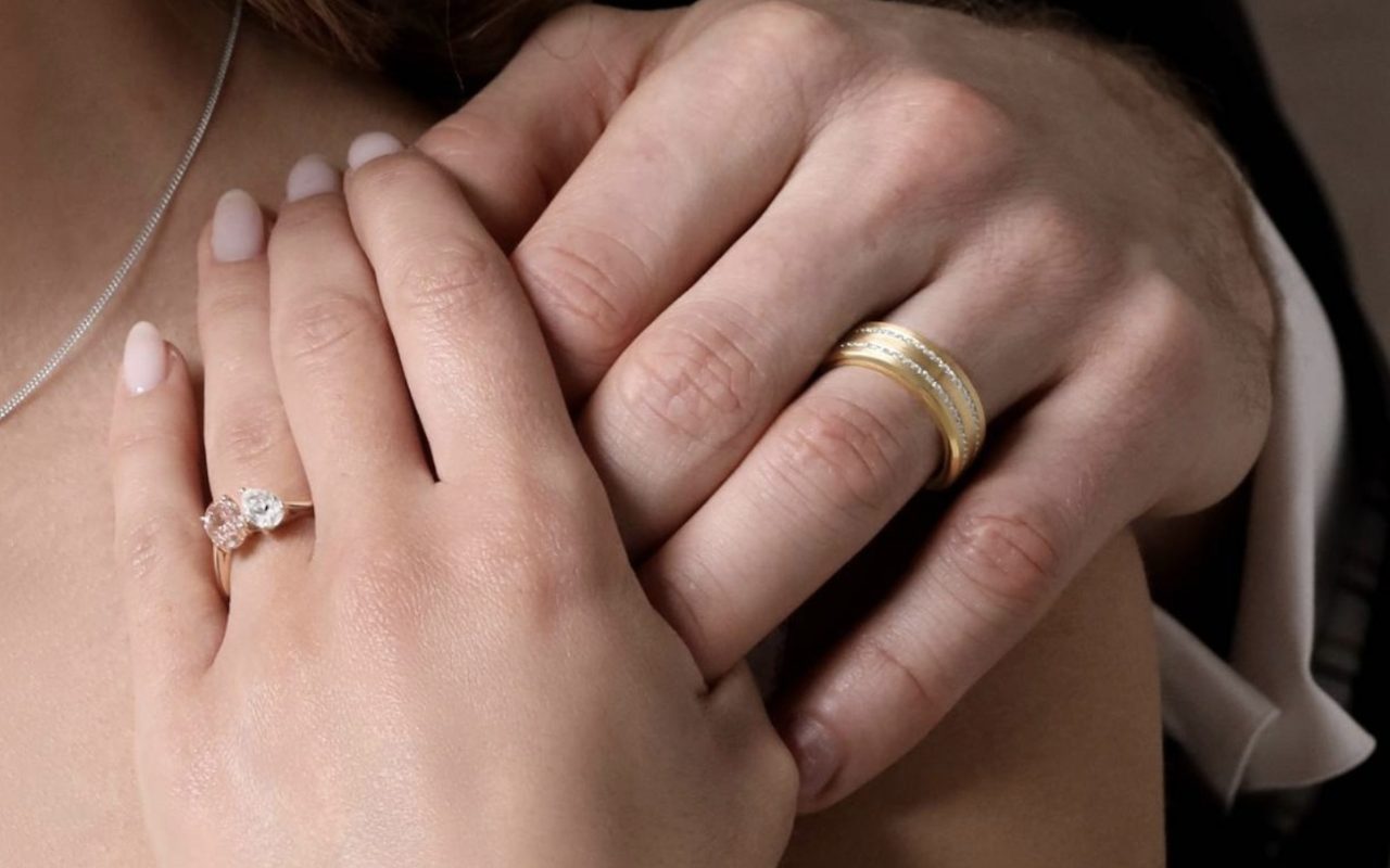 A diamond solitaire engagement ring on one hand and an 18ct gold wedding band on the other, a Jago Jewellers commission.